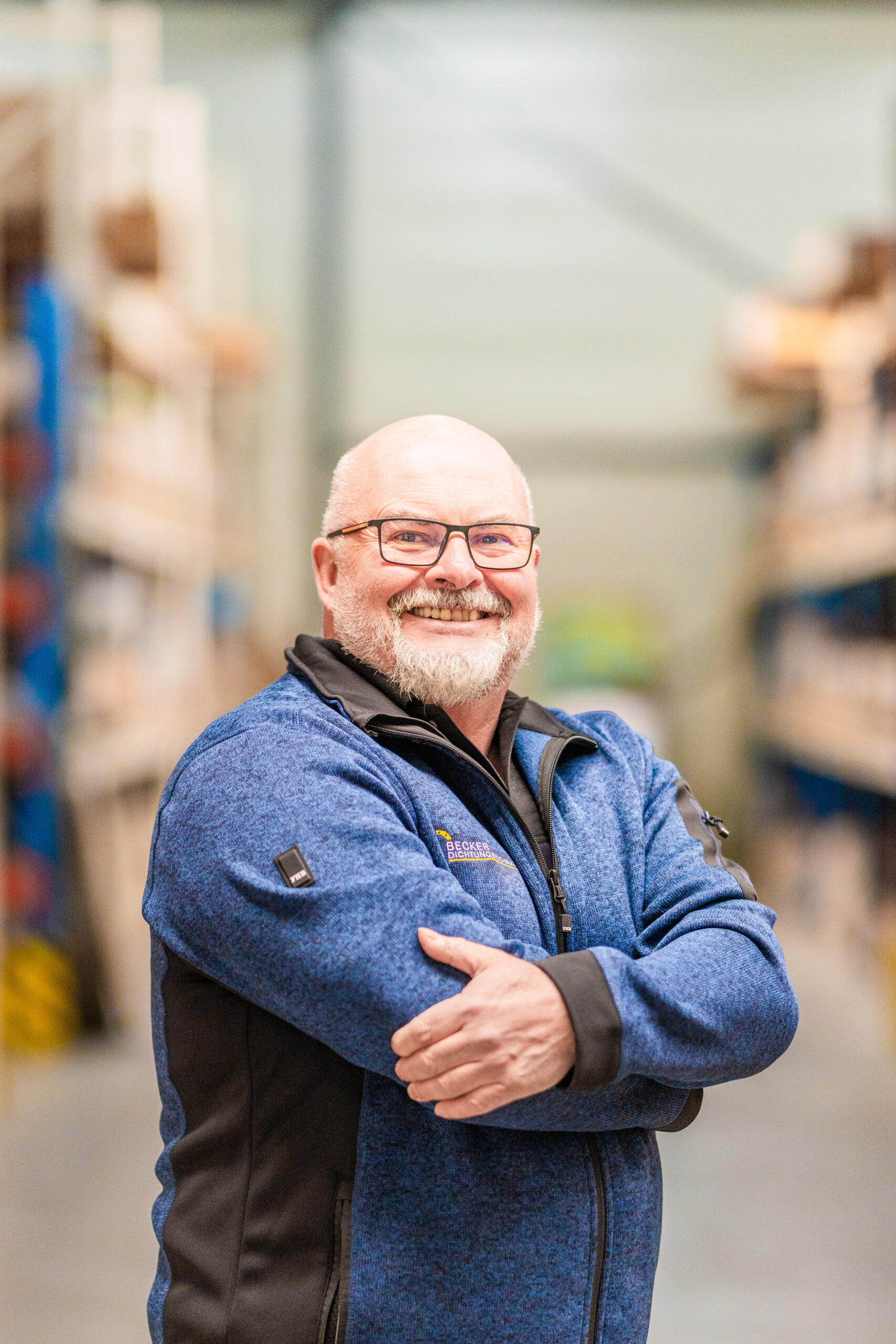 Smiling bald man with glasses and a blue jacket stands in a warehouse aisle with arms crossed.