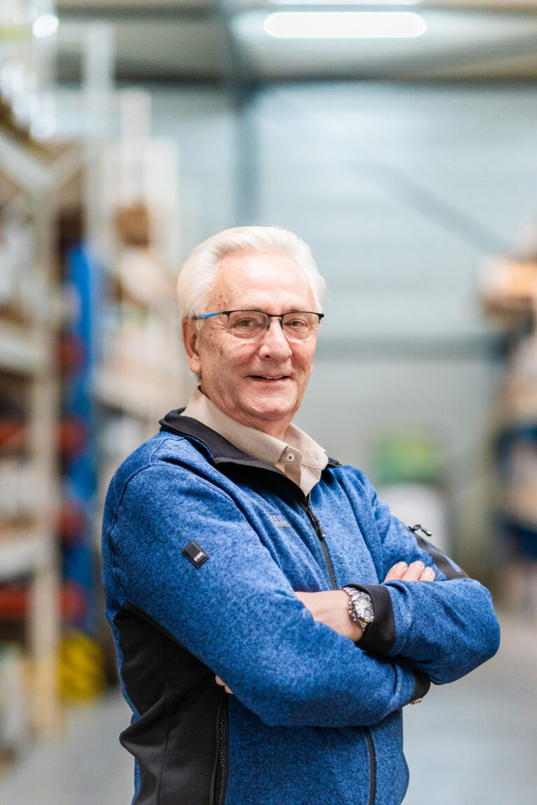 Older man with white hair and glasses, smiling and standing with arms crossed in a warehouse setting.