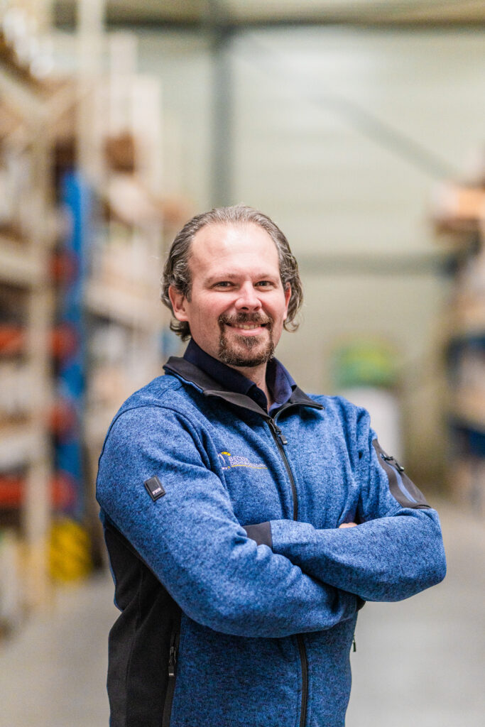 Man in blue jacket standing with arms crossed in a warehouse, smiling at the camera.