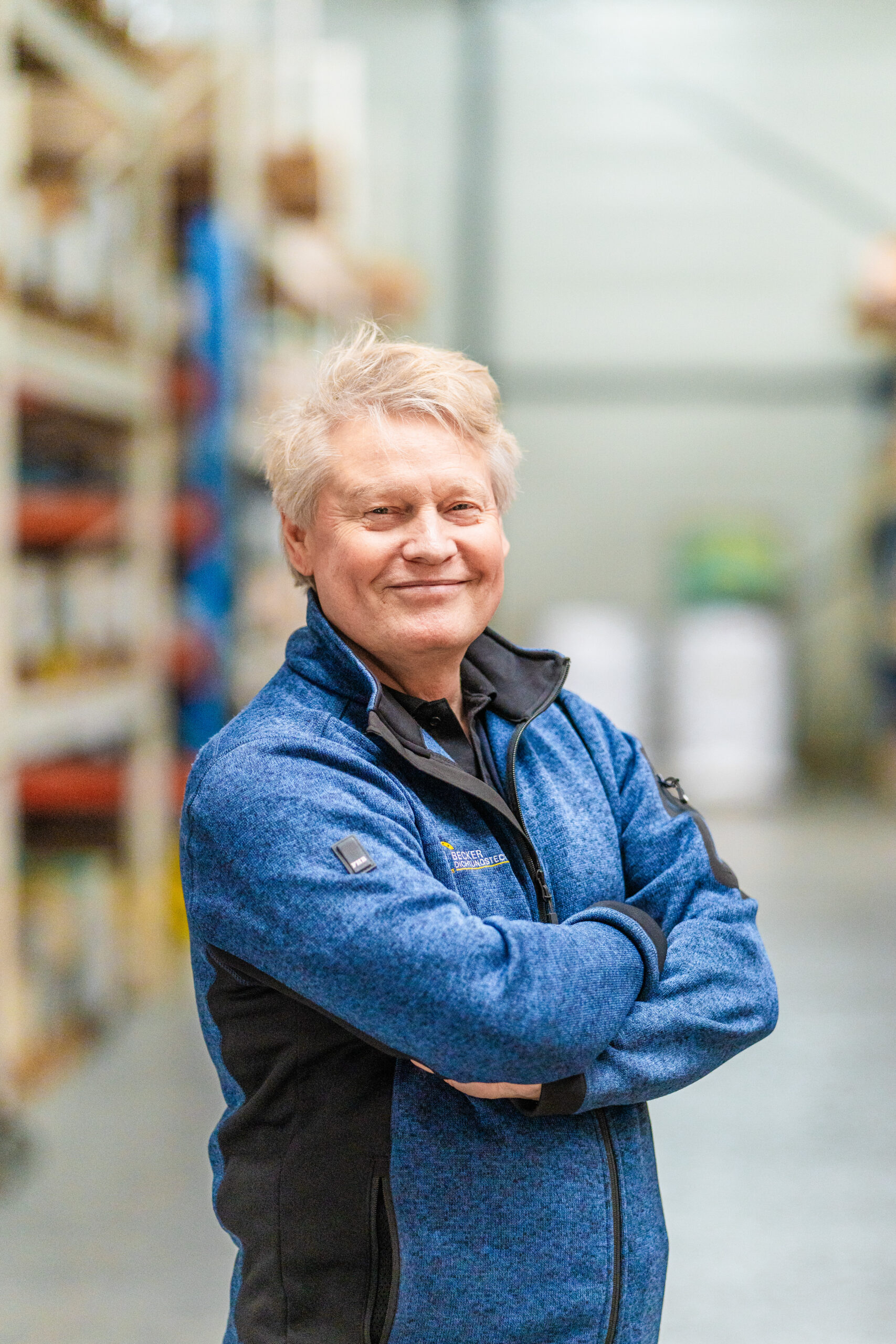 Older man with gray hair in a blue jacket stands smiling with arms crossed in a warehouse aisle.