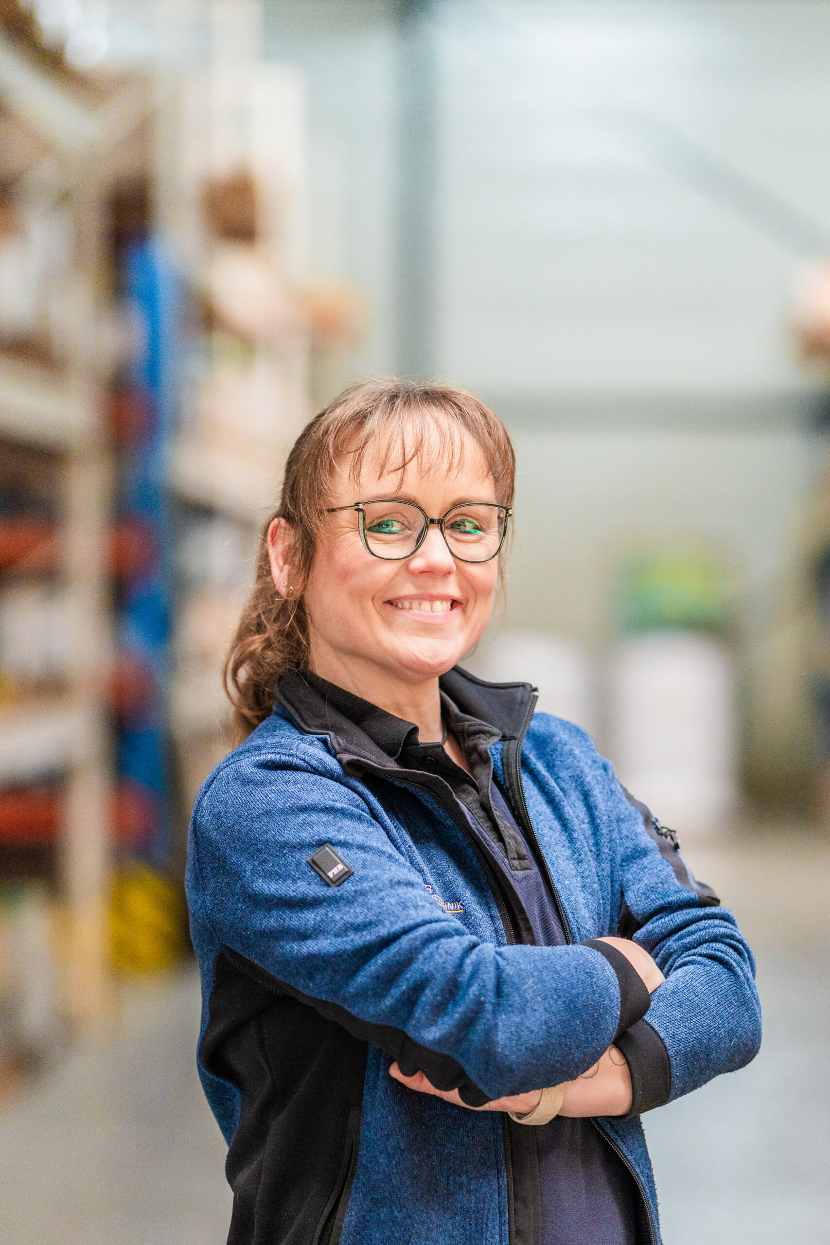 A woman with glasses and a blue jacket smiles, standing with arms crossed in a warehouse aisle.