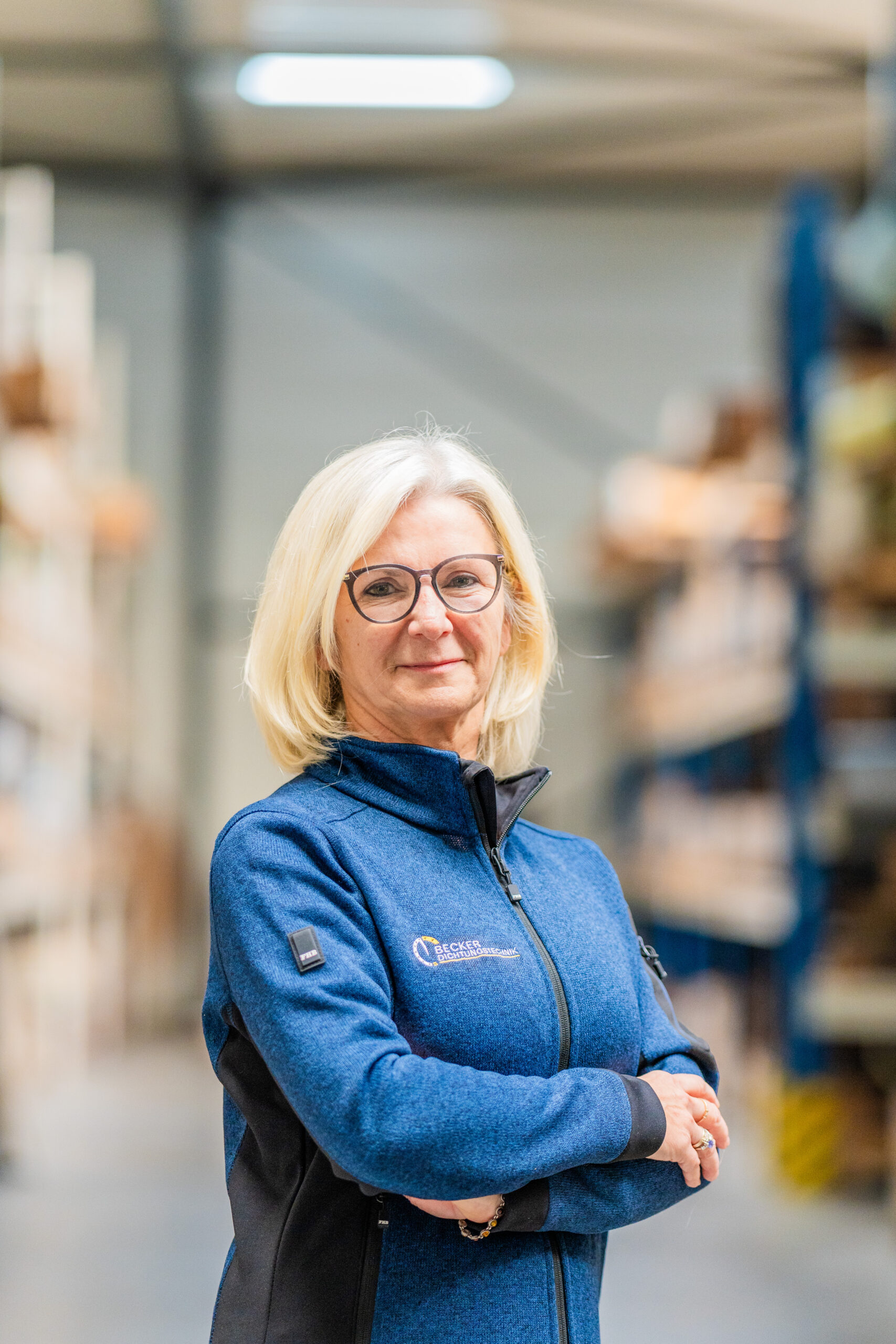 Confident woman with blonde hair and glasses, wearing a blue jacket, standing in a warehouse aisle.
