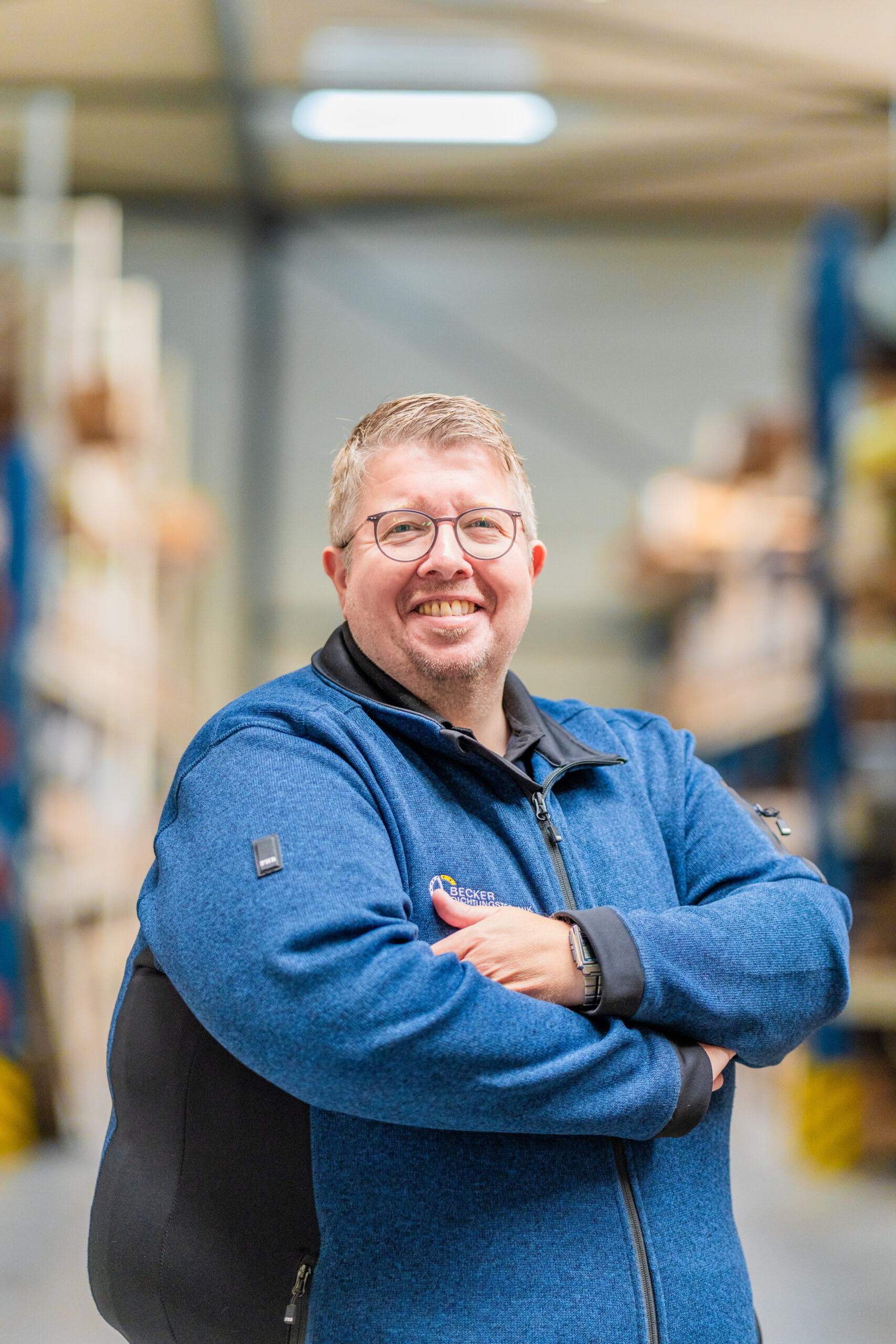 Smiling man with glasses and blue jacket stands in a warehouse with arms crossed.