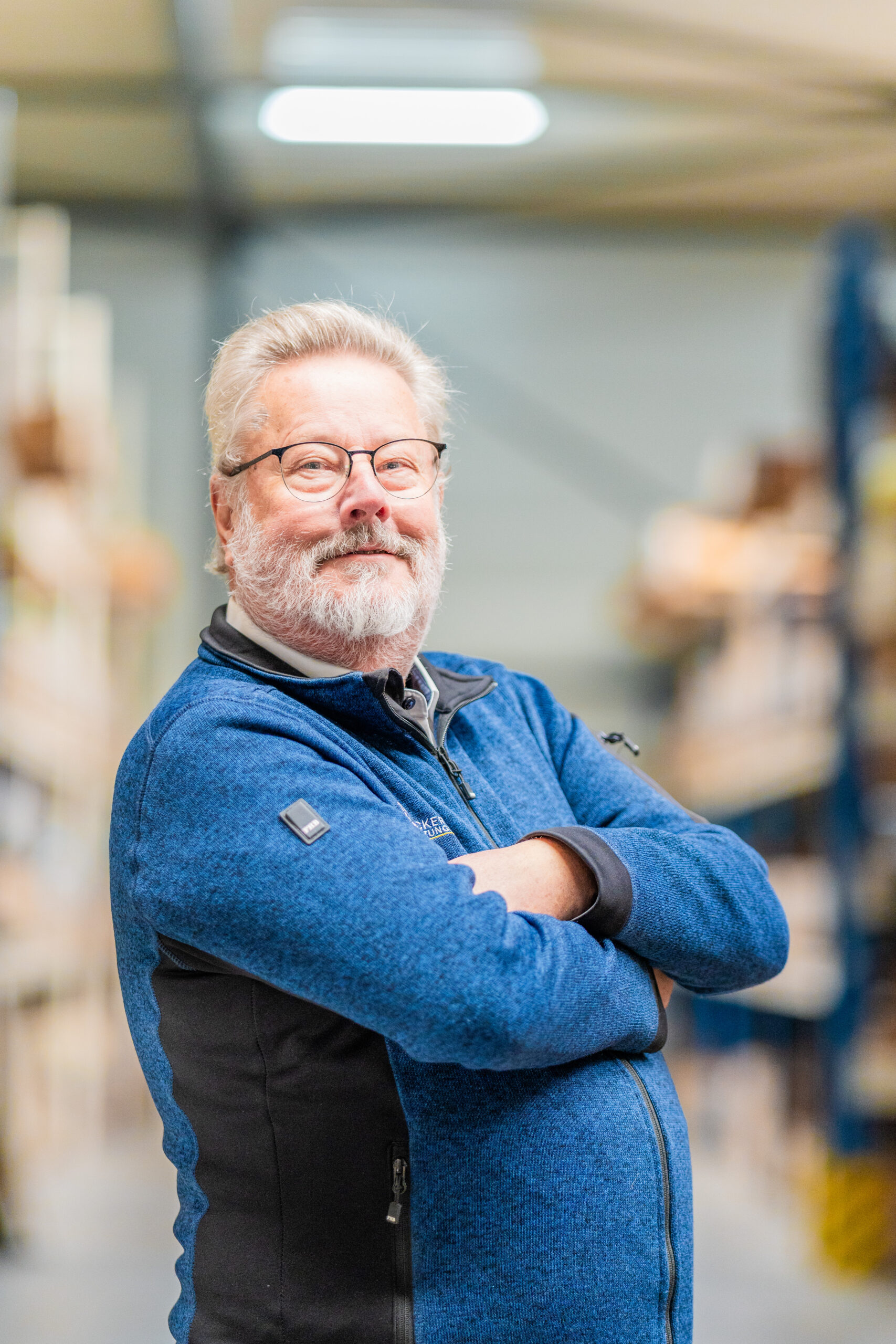 Older man with glasses and a blue jacket stands confidently with arms crossed in a warehouse.