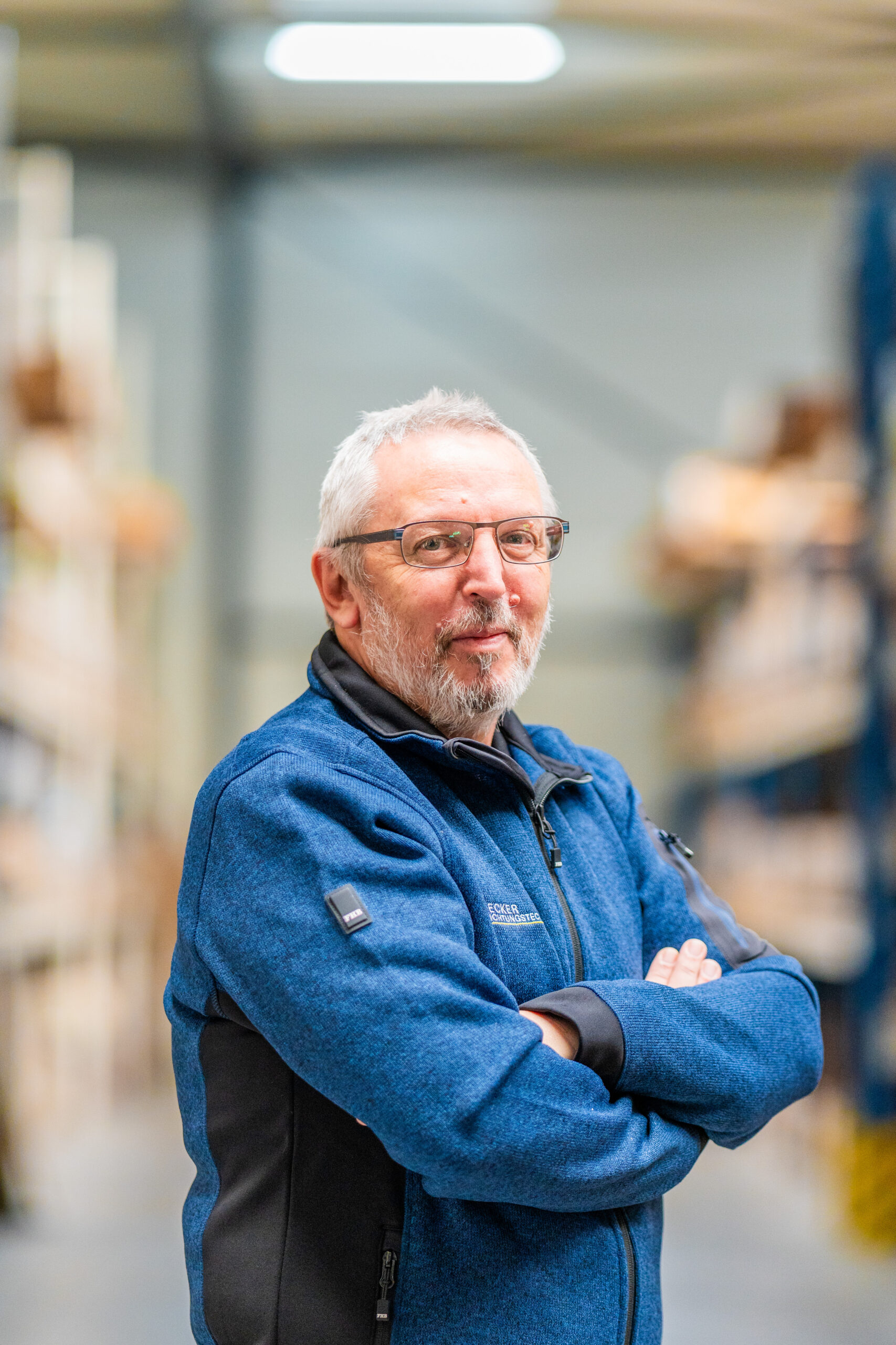 An older man with glasses and a blue jacket stands with arms crossed in a warehouse aisle.