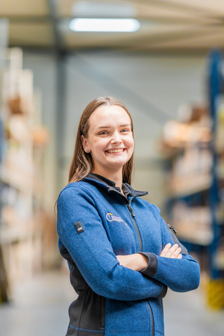 Smiling woman in a blue jacket stands with arms crossed in a warehouse aisle.