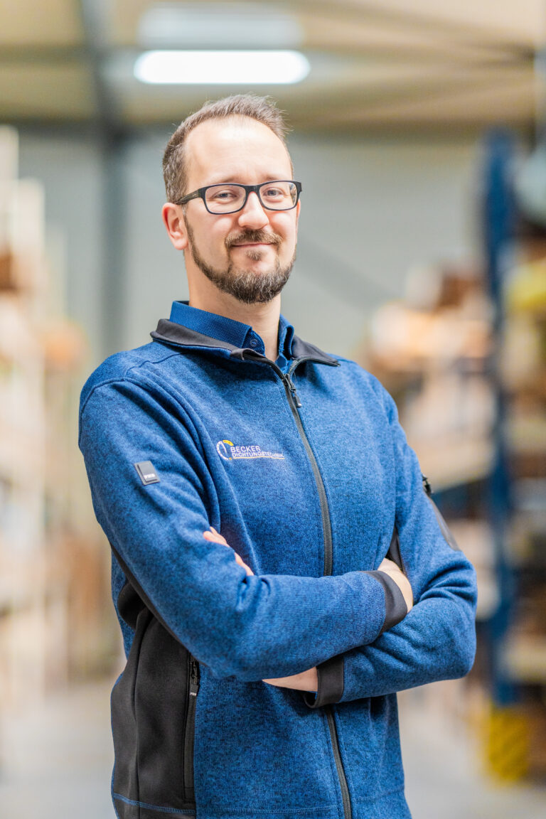Man with glasses and a beard, wearing a blue jacket, standing in a warehouse with arms crossed, smiling.