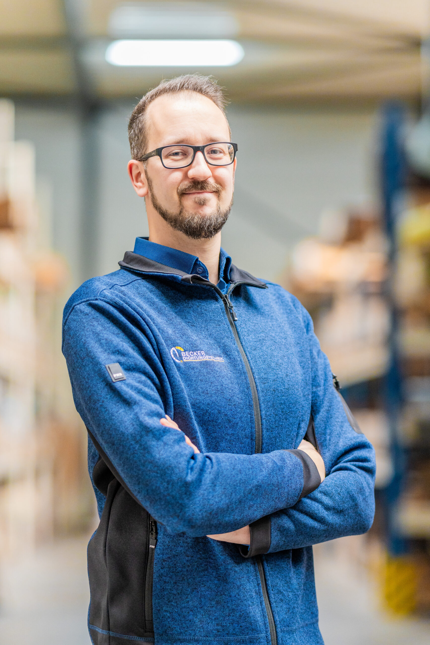 Man with glasses and a beard, wearing a blue jacket, standing in a warehouse with arms crossed, smiling.