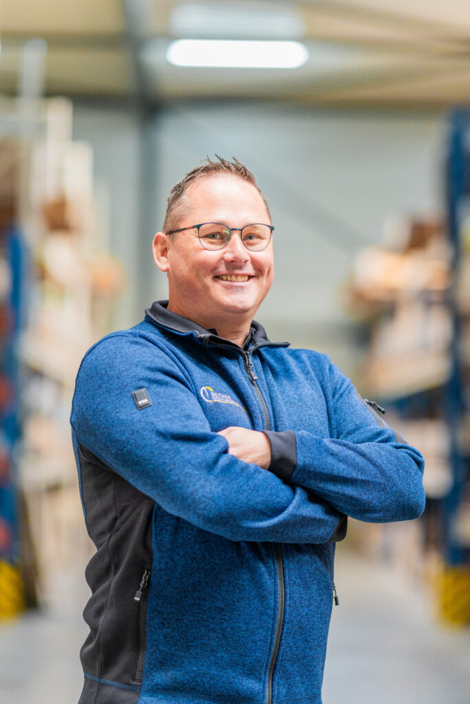 Smiling man in glasses and blue jacket stands with arms crossed in a warehouse aisle.