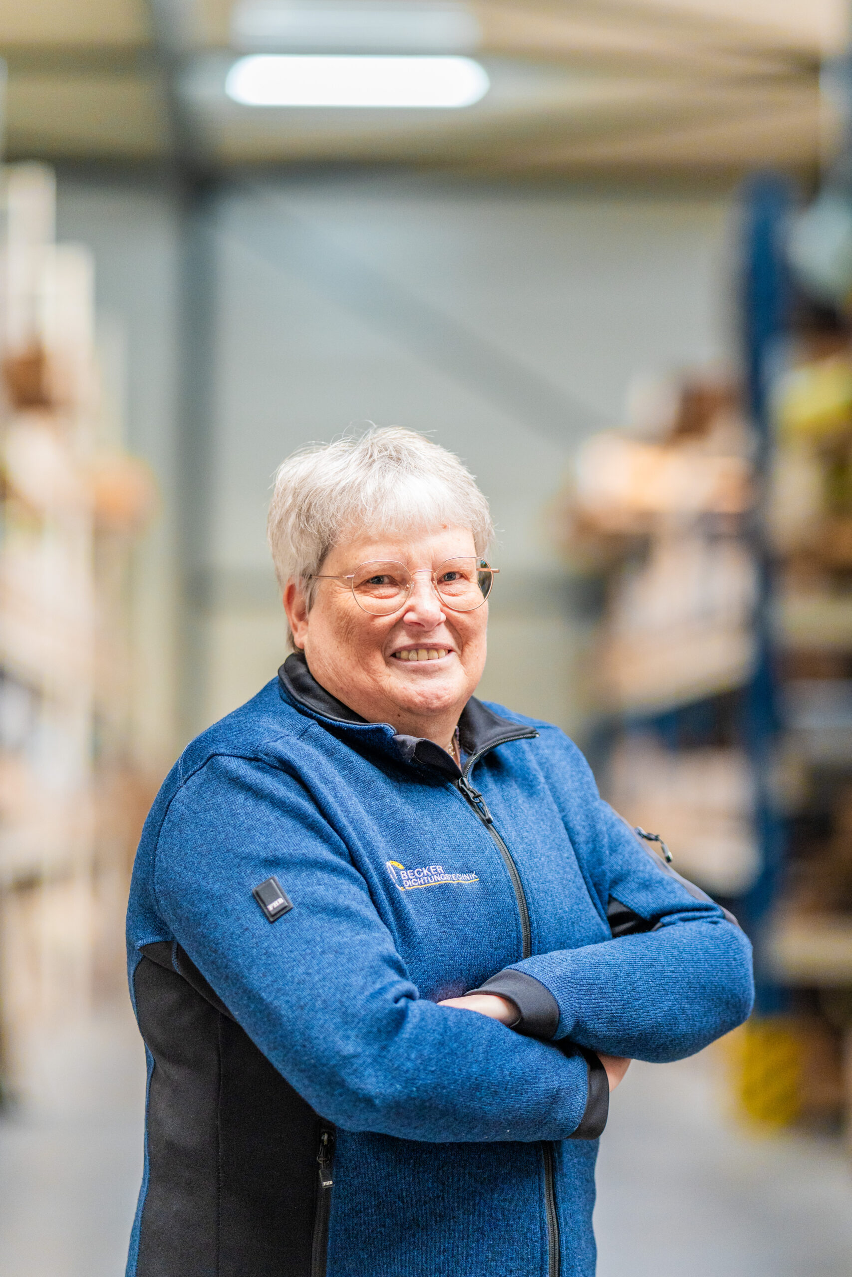 Smiling older woman with gray hair and glasses, standing with arms crossed in a warehouse aisle.