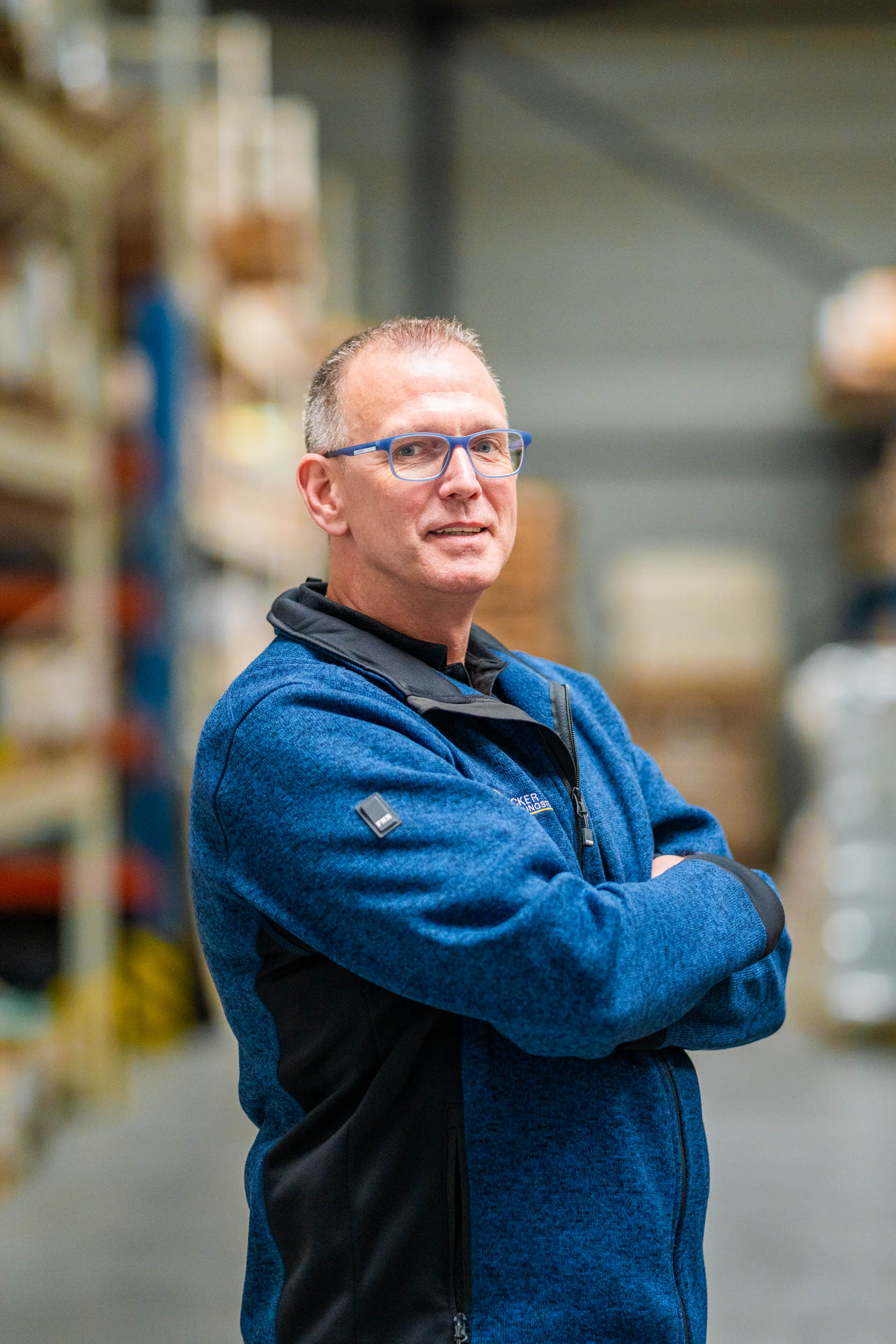 Man in a blue jacket with glasses standing in a warehouse, arms crossed, looking at the camera.