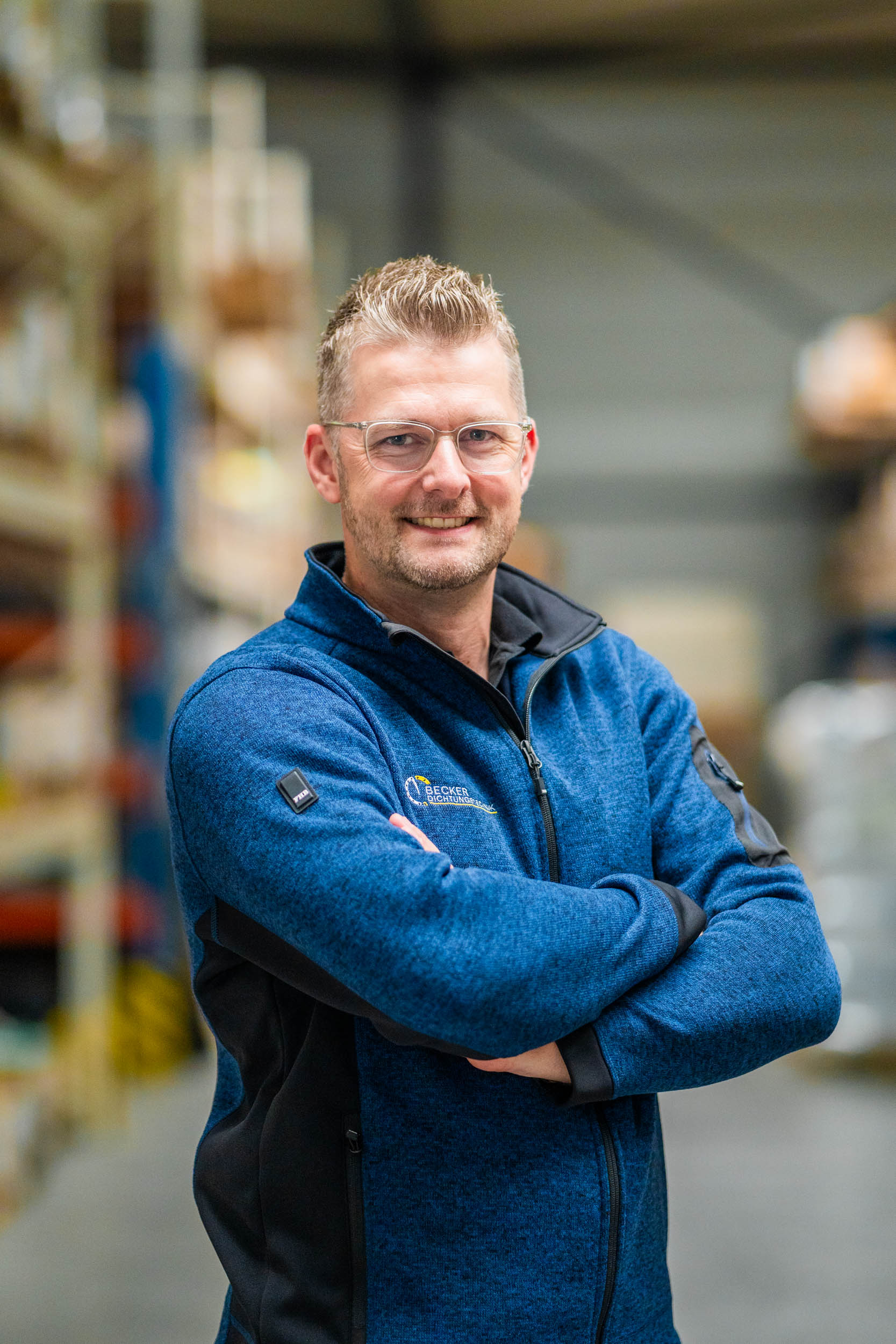 Smiling man with glasses and blue jacket stands with arms crossed in a warehouse aisle.