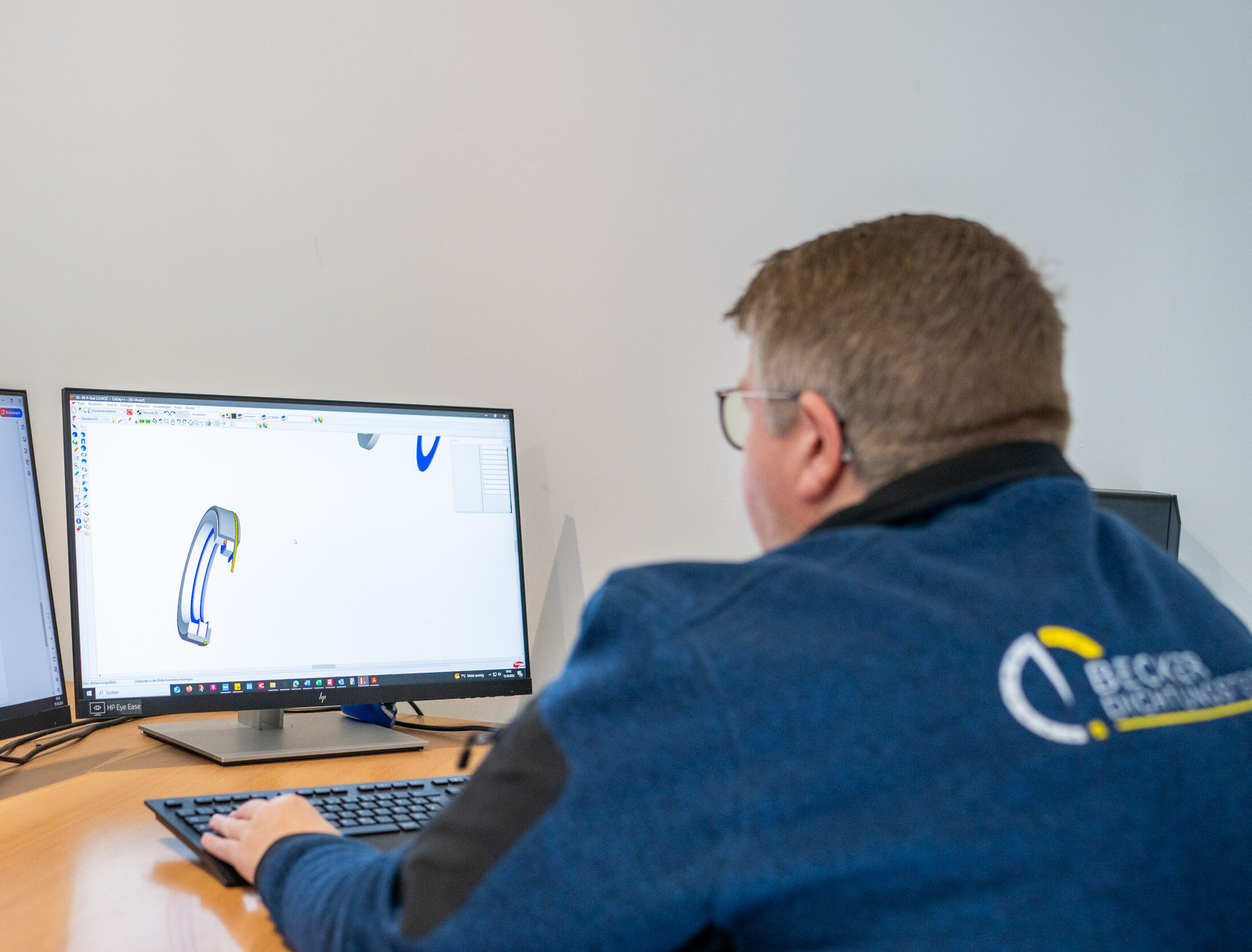 Person working on a 3D design on a computer, with technical drawings on a second monitor.