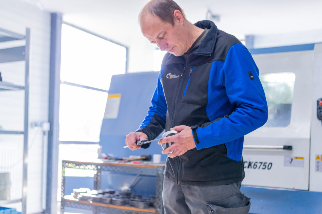Man in a workshop using a caliper to measure a metal object near industrial equipment.