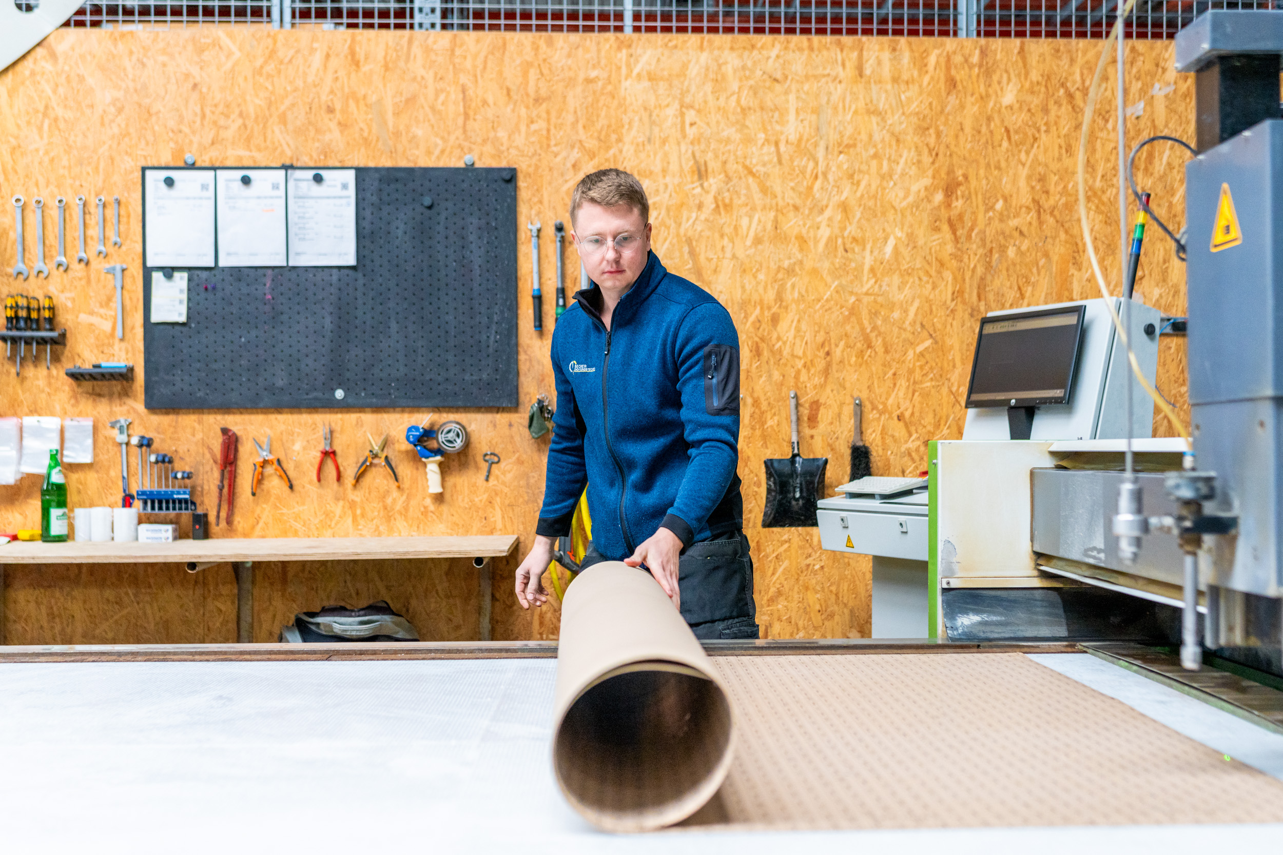 Man in a workshop rolling a large cardboard tube on a table near industrial equipment and tools.