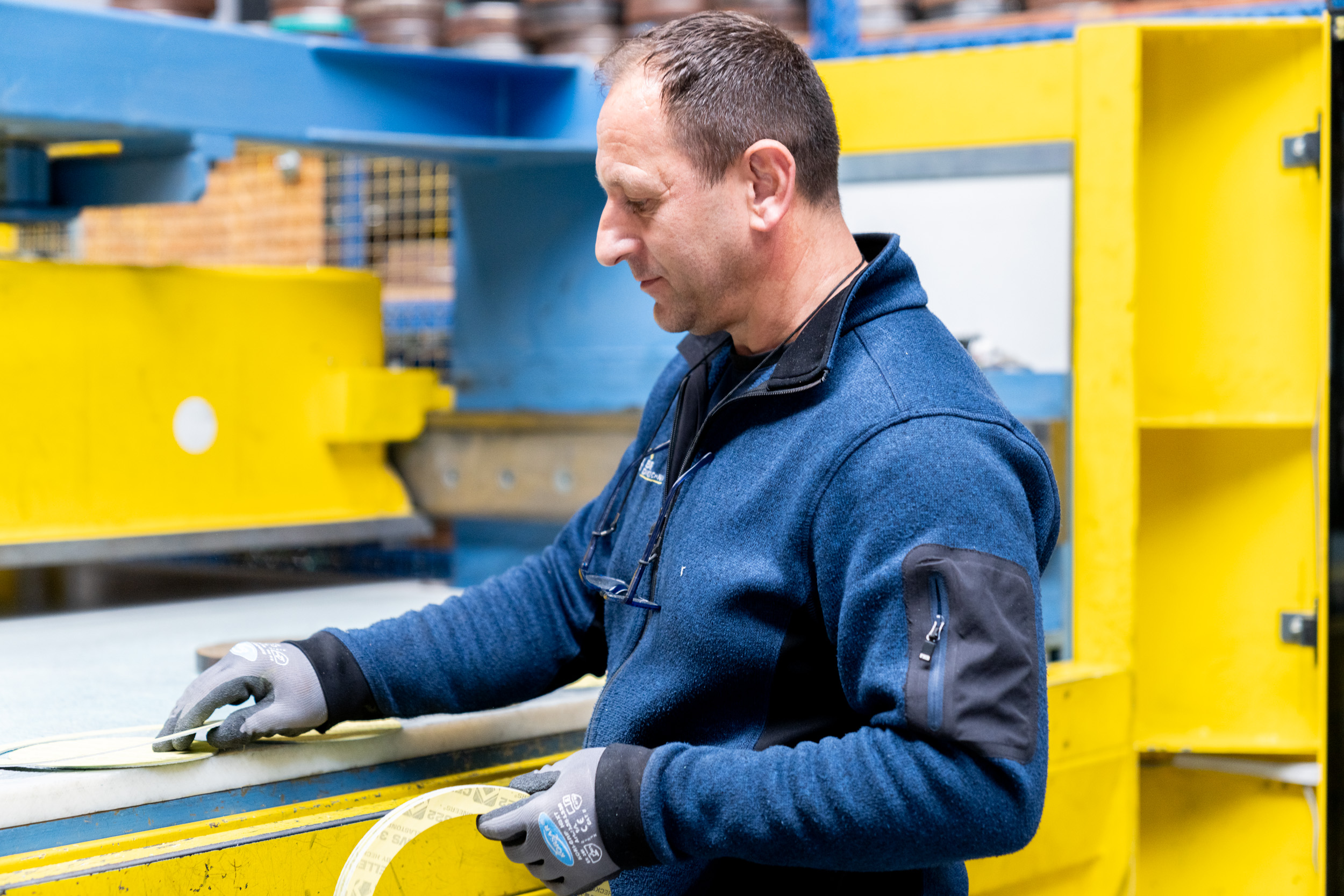Man wearing gloves measures material with tape in a brightly lit industrial setting.