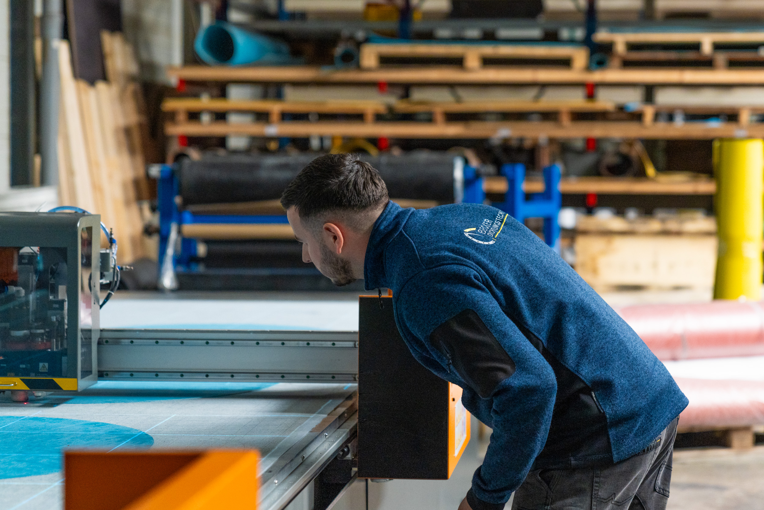 A man in a blue jacket operates and closely inspects a large cutting machine in a workshop.