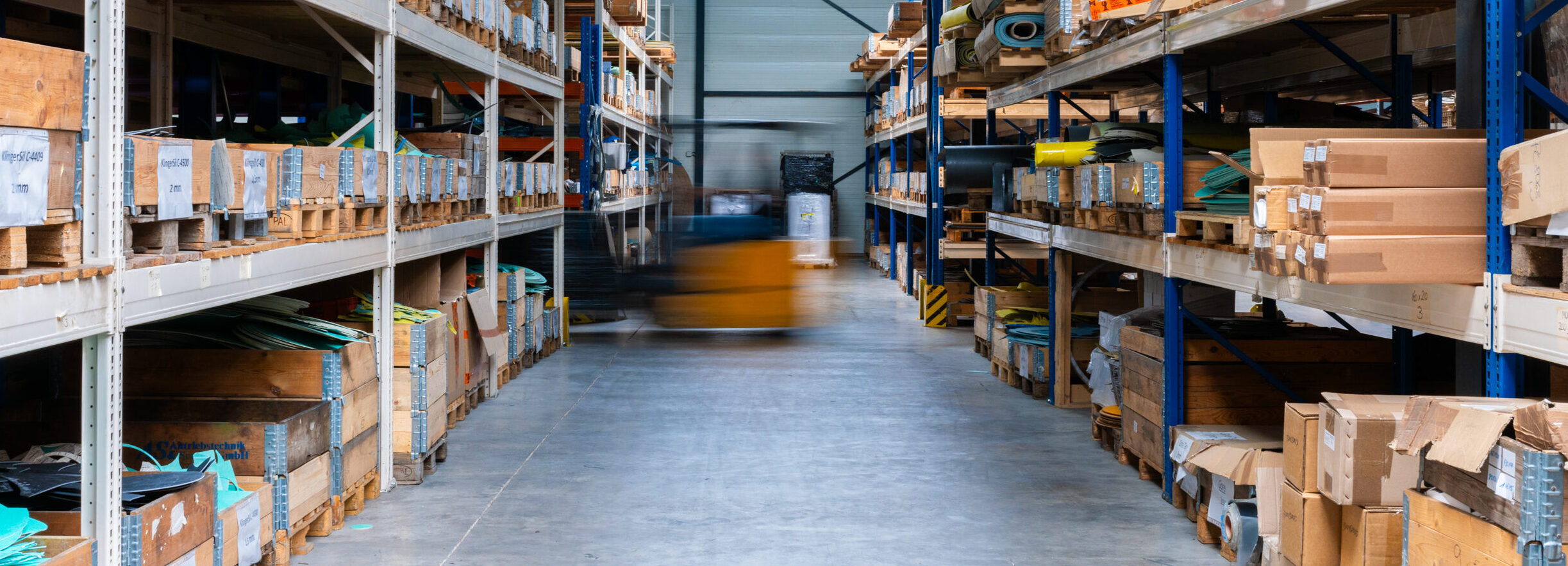Warehouse aisle with shelves storing boxes and materials, and a blurred forklift moving in the center.