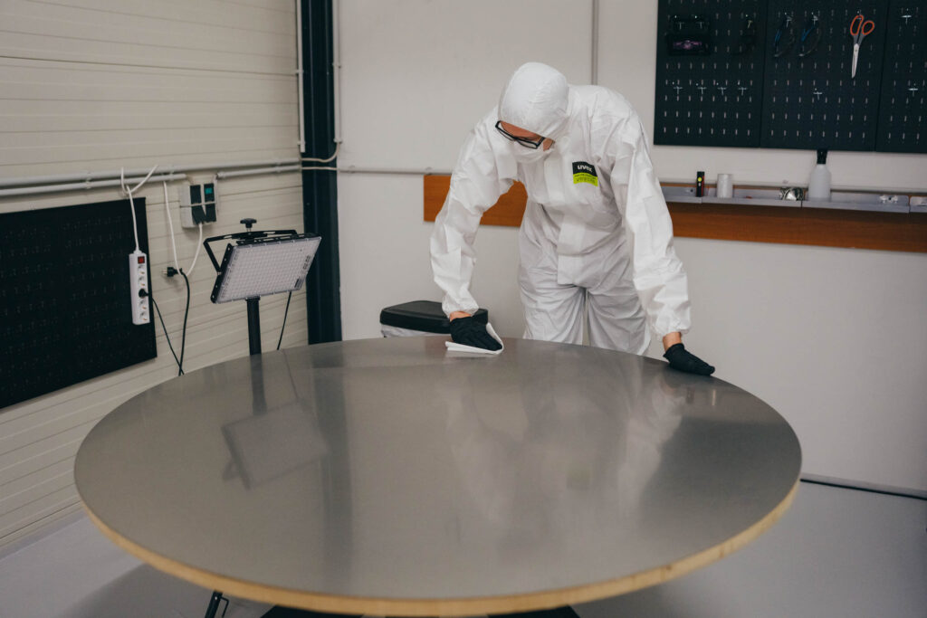 Person in protective suit cleaning a large round metal table in a laboratory or cleanroom setting.