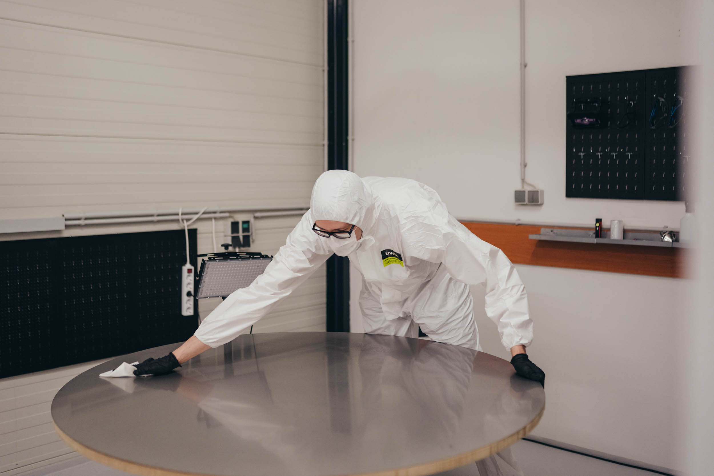Person in protective suit and gloves cleaning a round metal table in a laboratory or sterile environment.