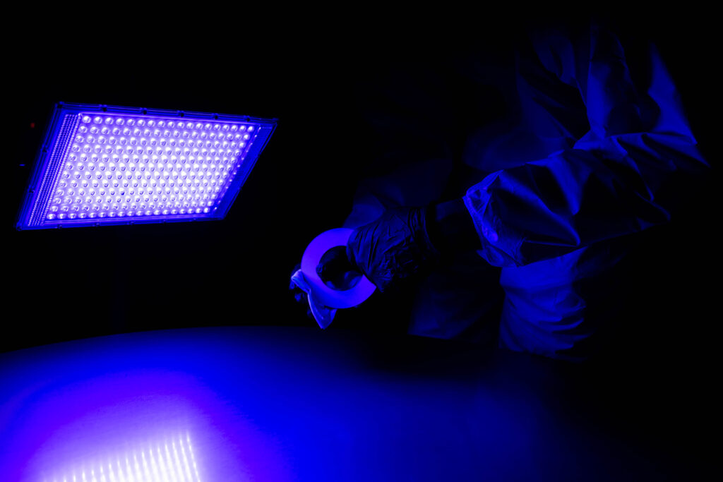 A person in gloves holds tape under a bright blue UV light, illuminating a smooth surface.